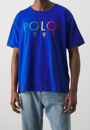 Uomo con maglietta blu con scritta colorata "POLO 992" e jeans azzurri chiari, ritagliato al collo e a metà coscia, in piedi davanti a uno sfondo semplice.