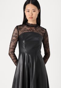Robe noire avec un buste ajusté, des manches longues en dentelle et une jupe évasée et lisse. Les détails en dentelle autour du décolleté ajoutent de la texture.
