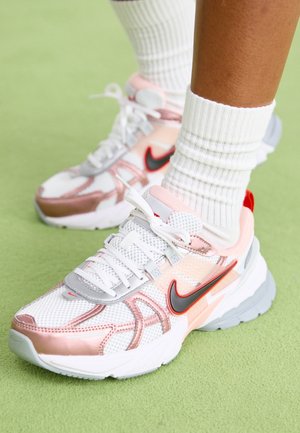 Scarpe da ginnastica Nike bianche e rosa con loghi swoosh rossi e neri, indossate con calzini bianchi a coste, su superficie verde.