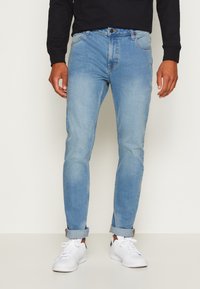 Világoskék farmernadrág, slim fit szabással, gombos záródással és feltekerhető szárakkal, fehér sportcipővel viselve.