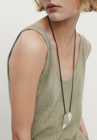 Green knitted tank top with a relaxed fit, featuring textured fabric and wide straps. A long black cord necklace with a silver pendant is worn.