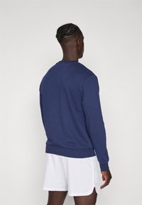 Man met gevlochten haar, gekleed in een marineblauwe langemouwen shirt en witte sportshorts, gezien van achteren tegen een effen achtergrond.