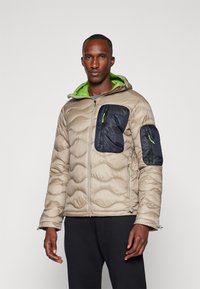 Padded jacket in beige with dark blue accent pockets. Features a green-lined hood, textured fabric, and adjustable cuffs for comfort.