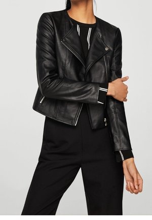 Frau trägt eine enganliegende schwarze Lederjacke über einem schwarzen Outfit mit weißen vertikalen Streifen an den Ärmeln und steht mit einem Arm über den anderen verschränkt.