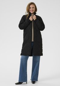 Manteau long matelassé noir avec un col haut, poches zippées, poignets ajustés et coupe droite, associé à un jean bleu à jambes larges et des chaussures noires.