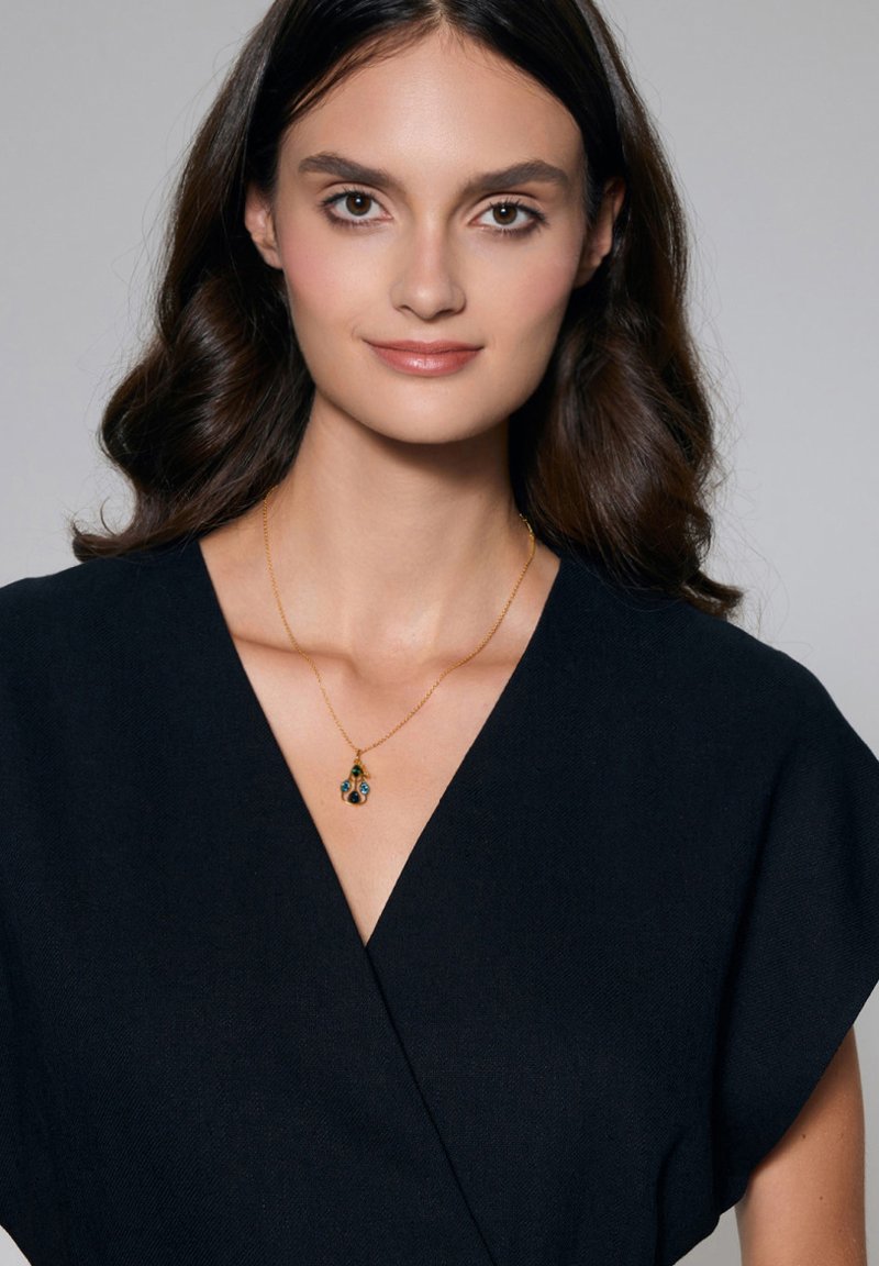 Złoty naszyjnik z zawieszką w niebiesko-zielonych akcentach na cienkim łańcuszku, noszony na ciemnej, fakturowanej sukience. Prosty i elegancki design.