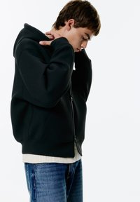 Felpa nera da uomo con zip, realizzata in tessuto morbido, con cappuccio e orlo a costine. Indossata sopra un maglione chiaro e jeans blu.
