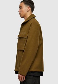Veste en mélange de laine vert olive avec un design à col, poche avant et fermeture zippée, présentant une silhouette carrée et une texture douce.