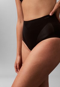 Kvinde iført sort Calvin Klein undertøj med høj talje, der viser overkroppen og det øverste af lårene mod en ensartet grå baggrund.