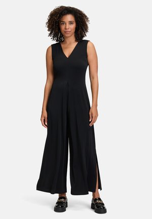 Zwarte mouwloze jumpsuit met een V-hals, wijde pijpen en zijsplitten, gemaakt van gladde stof, gecombineerd met chunky zwarte schoenen met gouden hardware.