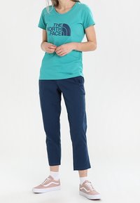 T-shirt turchese con logo blu navy, abbinato a pantaloni blu scuro e sneakers rosa chiaro. I pantaloni sono corti; le sneakers hanno suole bianche.