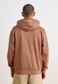 Sweat à capuche marron avec une texture douce, doté d'une grande capuche et du lettrage « JORDAN » sur le haut du dos. Coupe décontractée avec ourlet et poignets côtelés.