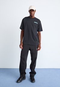 Sort oversized t-shirt med turkis logo, parret med sorte atletikbukser med sidelommer. Båret med en lysefarvet kasket og sorte sneakers.