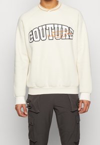 White crewneck sweatshirt with "COUTURE" graphic in black and orange. Features ribbed collar and cuffs, with a relaxed fit.