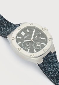 Montre-bracelet Guess ton argent avec cadran texturé noir, trois sous-cadrans, marqueurs blancs, aiguilles argentées et bracelet en tissu bleu à motif noir.