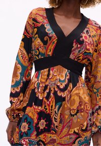 Robe à manches longues et décolleté en V, de couleur noire avec un imprimé floral jaune moutarde et rouge. Présente une taille élastique noire uni et un détail de jupe plissée.