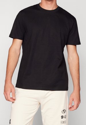 Schwarzes Baumwoll-T-Shirt mit kurzen Ärmeln, Rundhalsausschnitt und lockerem Schnitt, präsentiert an einem Modell. Kombiniert mit hellen Hosen.