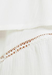 Bílá látka s texturovaným, zmačkaným vzhledem. Dekorativní lem má hnědý vzor a bílé švy podél okraje.