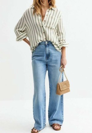 Vrouw in een lichtgroen en witte gestreepte blouse, hooggetailleerde blauwe jeans, bruine sandalen en een beige geweven handtas.