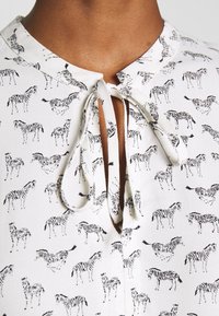 Vit skjorta med svart zebra-mönster, knytkrage, och kort placket. Slät tygkänsla, med spridda zebraillustrationer.