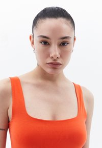 Top de tirantes anchos y escote cuadrado en color naranja, hecho de una tela suave y elástica. El tono de piel es claro con pecas.