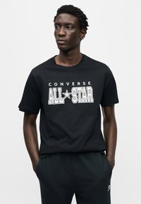 Μαύρο βαμβακερό T-shirt με στρογγυλή λαιμόκοψη, με μεγάλο λευκό κείμενο "CONVERSE ALL STAR" στο στήθος. Απλός, καθημερινός σχεδιασμός.