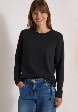 Femme souriante portant un t-shirt noir uni à manches longues et un jean bleu, les mains partiellement dans les poches, sur un fond neutre.
