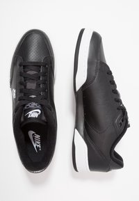 Zapatillas Nike negras con parte superior de cuero suave, laterales perforados y suela de goma blanca. Presenta cordones negros y un forro interior de la marca.