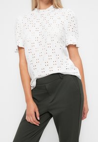 Vit ögonbladsblus med puffärmar, rund halsringning, som har utskurna mönster. Kombinerad med mörkgröna skräddarsydda byxor.