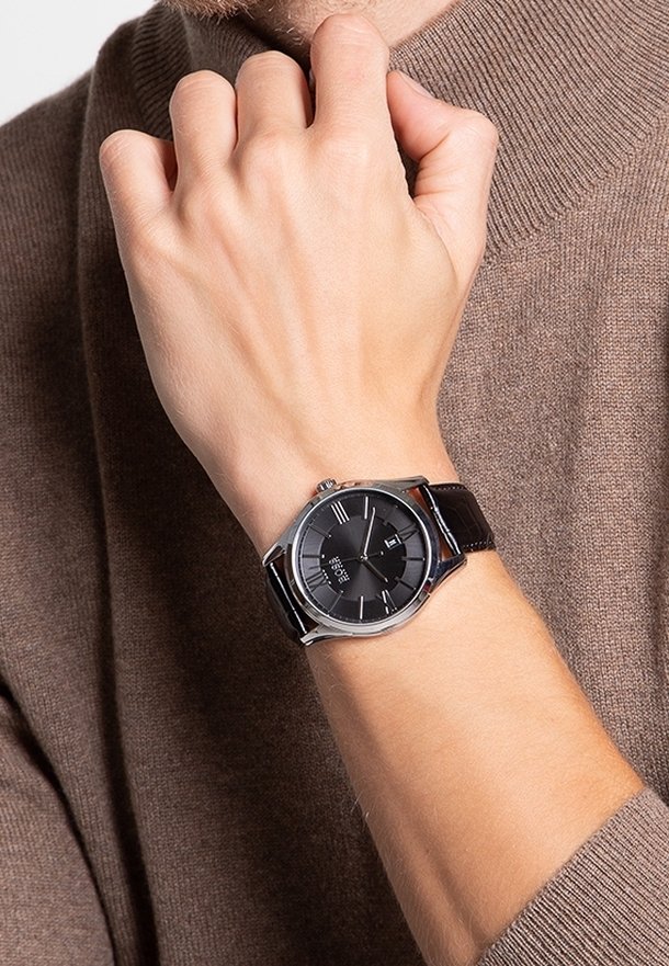 Poignet masculin portant une montre analogique avec un bracelet en cuir noir, un cadran noir et un boîtier argenté, sur un pull en maille marron.