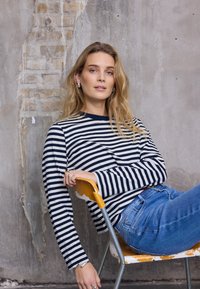 Pika varrukaga tumesiniste ja valgete triipudega top koos siniste teksadega, istub kulunud oranži ja metallist tooli peal tekstuurse seina taustal.