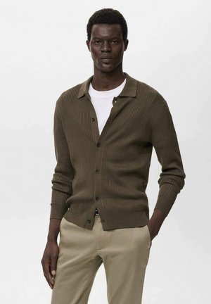 Man wearing olive green ribbed buttoned cardigan over white T-shirt and beige trousers, standing with one hand in pocket against plain background.