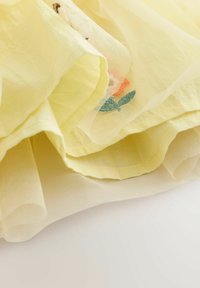 Tissu superposé de couleur jaune clair avec une texture légère et transparente, présentant des broderies florales orange et vert. Apparence douce et délicate.