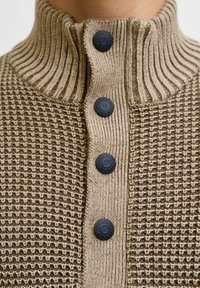 Beżowy sweter dzianinowy z prążkowanym, wysokim kołnierzem i czterema czarnymi guzikami z wygrawerowanym napisem "JACK & JONES EST. 1990" na zapięciu.
