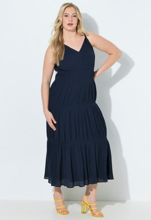 Lyshåret kvinde iført en ærmeløs navyblå midikjole og gule, hælede sandaler, poserer med den ene hånd på hoften foran en hvid baggrund.