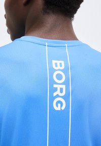 Κοντινό πλάνο του άνω μέρους της πλάτης ενός ατόμου που φοράει μπλε υφασμάτινο πουκάμισο με λευκές κάθετες γραμμές και τη λέξη «BORG» τυπωμένη κάθετα.