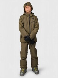 Παιδί με καφέ εξοπλισμό σκι και σκουφάκι, φορώντας μαύρα γάντια και ασημένια μποτάκια σκι, που στέκεται μπροστά σε γκρίζο φόντο.