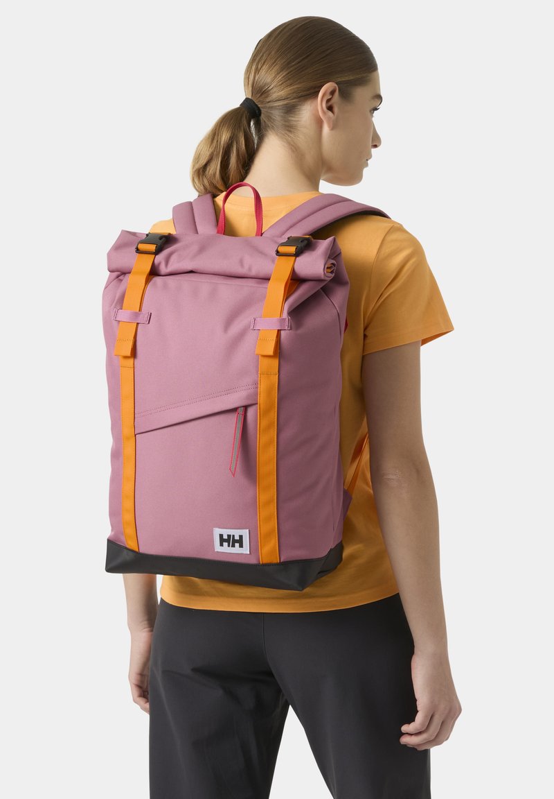 Mujer con cabello castaño recogido en una coleta, vistiendo una camiseta naranja y pantalones negros, llevando una mochila enrollable morada y naranja con el logo HH.