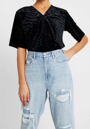 Femme portant un haut noir texturé à col en V avec des manches jusqu'au coude et un jean déchiré taille haute bleu clair.