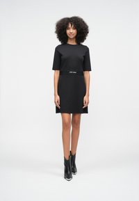 Frau mit lockigem Haar, die ein schwarzes, kurzärmeliges Kleid und schwarze Stiefeletten trägt und vor einem schlichten weißen Hintergrund steht.