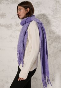 Écharpe à franges violettes en tissu doux, drapée autour du cou, associée à un pull en tricot couleur crème et à un pantalon noir.
