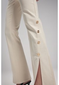 Pantaloni beige a palazzo con fessure laterali, dotati di dettagli con bottoni dorati lungo la cucitura, realizzati in un tessuto liscio e leggermente strutturato.