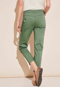 Pantalons verts coupés avec une coupe slim, dotés de deux poches arrière et d'une texture lisse. Portés avec des chaussures brunes à enfiler.