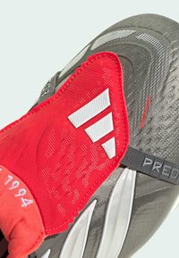 Grå og rød atletisk sko med en struktureret overflade, hvide detaljer og en tætsiddende pasform; inkluderer branding og en mærket rem.