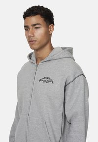 Felpa grigia con zip realizzata in tessuto morbido, dotata di tasca a marsupio, cappuccio regolabile e logo ricamato nero sul petto.