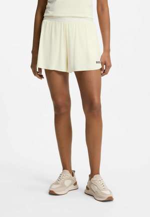 Person iført lysegule løse shorts med "BOSS" logo på venstre kant, kombineret med beige og hvide sneakers, stående på en hvid baggrund.
