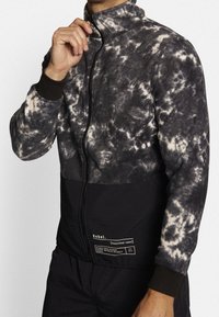 Giacca in pile con zip e motivo tie-dye in grigio e crema, con un pannello inferiore nero e polsini elasticizzati. Toppa con logo sul lato sinistro.