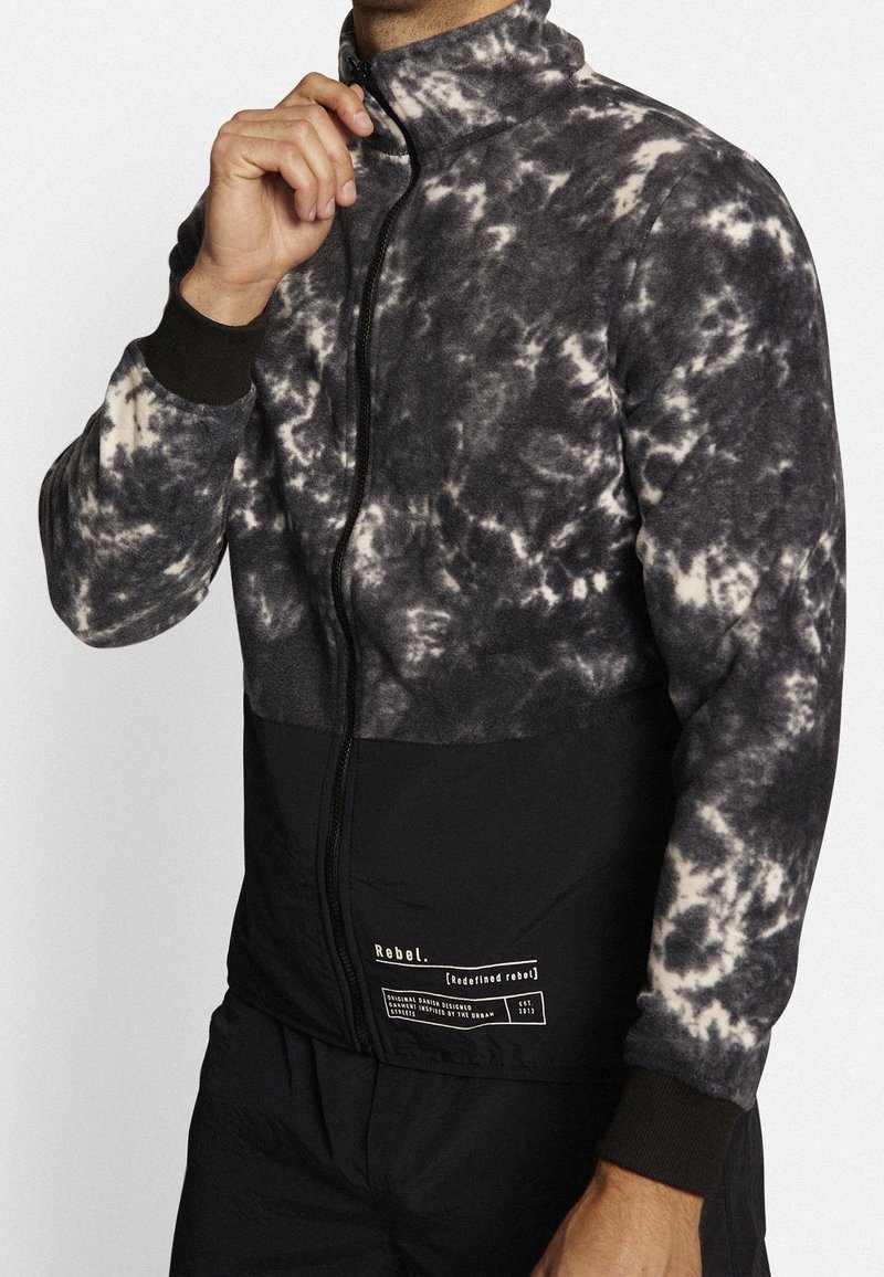 Giacca in pile con zip e motivo tie-dye in grigio e crema, con un pannello inferiore nero e polsini elasticizzati. Toppa con logo sul lato sinistro.