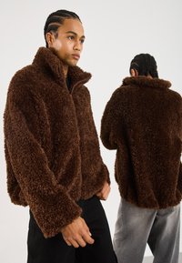 Man met gevlochten haar, die een bruine, fluffy, ritsjack modelleert, zowel van voren als van achteren, tegen een effen witte achtergrond.