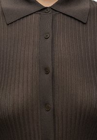 Gros plan d'une chemise en maille côtelée brun foncé avec un large col et quatre boutons noirs visibles sur le devant.